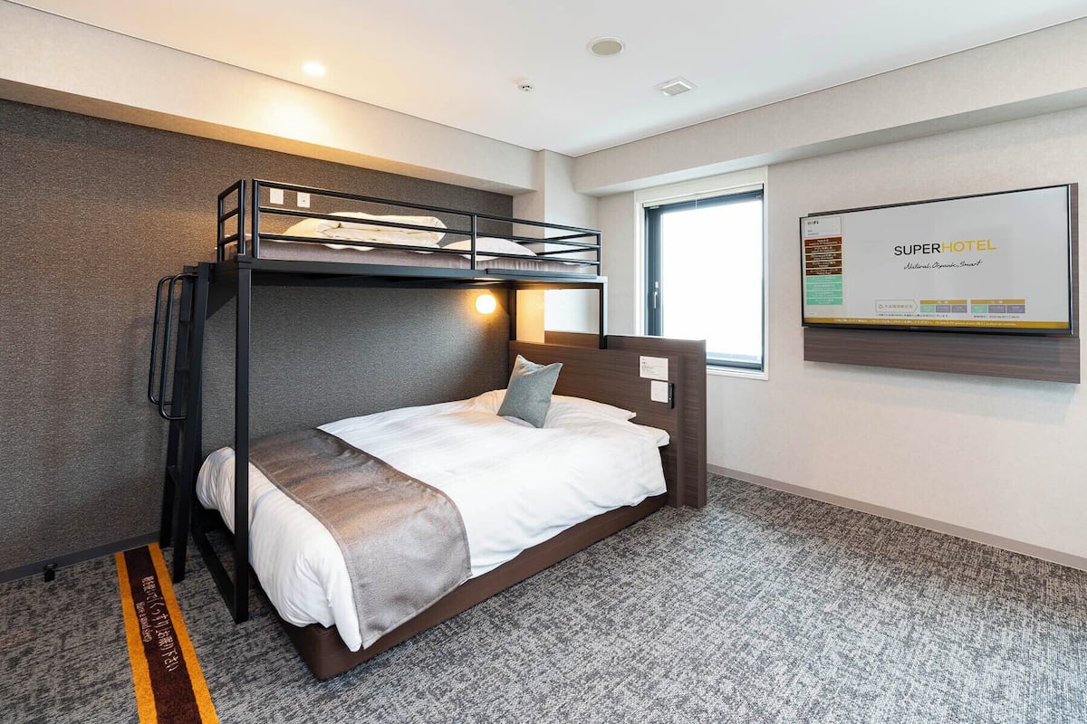Ein Schlafraum mit Etagenbetten, einem Fenster und einem an der Wand montierten Bildschirm, der 'SuperHotel' anzeigt.
