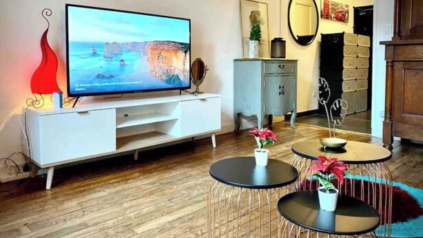 Ein modernes Wohnzimmer mit einem Flachbildfernseher auf einer weißen Unterhaltungsanlage, einem kleinen Schrank und zwei runden Tischen mit Topfpflanzen.