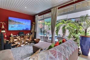 A modern living room with a large TV, comfortable seating, a view of a pool, and tropical plants. - Fare Nãhina - 2 chambres - Centre-Ville (Papeete)