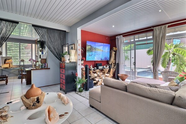 A modern living room with a sofa, a television, and a dining area with a table and decorative items. - Fare Nãhina - 2 chambres - Centre-Ville (Papeete)