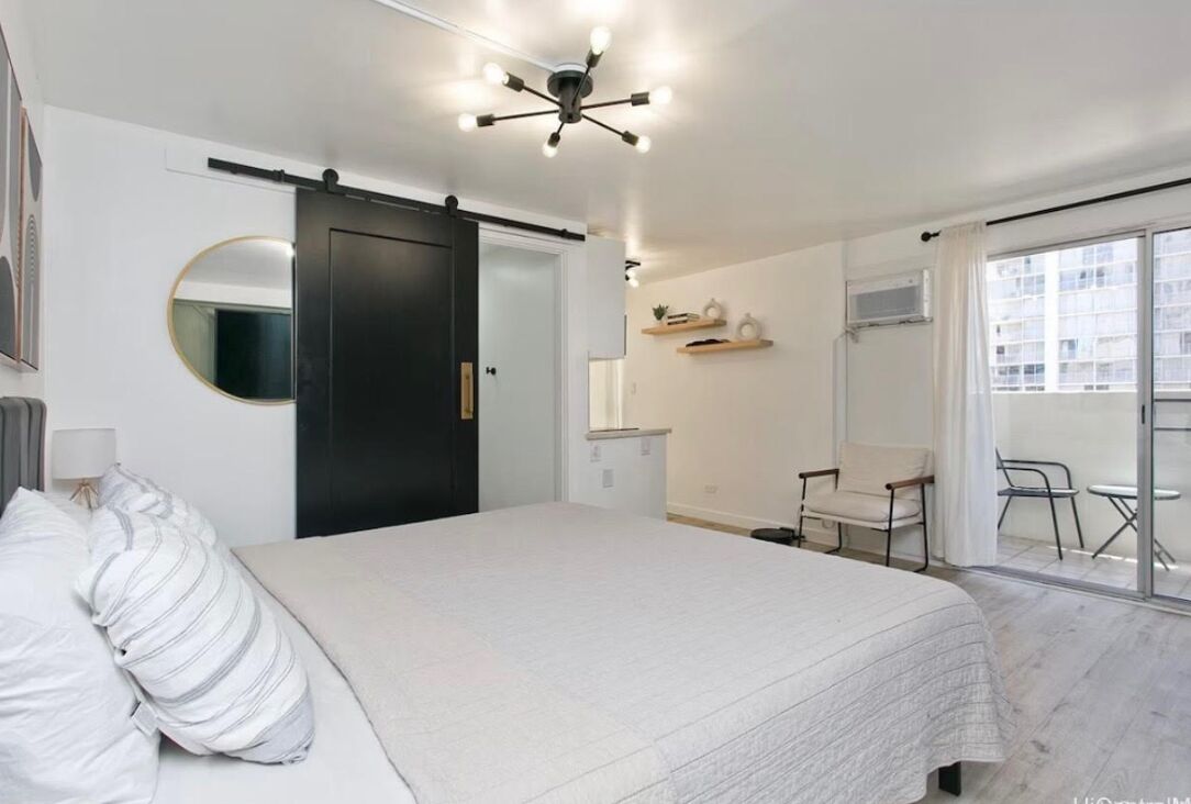 Un dormitorio con cama, una puerta corrediza, una mesita, una silla, un espejo y un balcón con vistas a edificios.