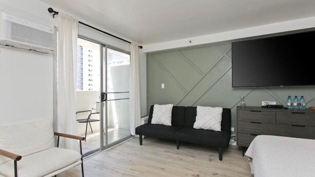 A modern hotel room with a sofa, a bed, a TV, a dresser, and a sliding glass door to the balcony. - Aloha Suite Near the Sea IV, 3 Block Walk to Beach (Honolulu)
