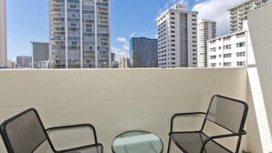 A balcony with two chairs and a small table, overlooking a cityscape with various buildings. - Aloha Suite Near the Sea IV, 3 Block Walk to Beach (Honolulu)