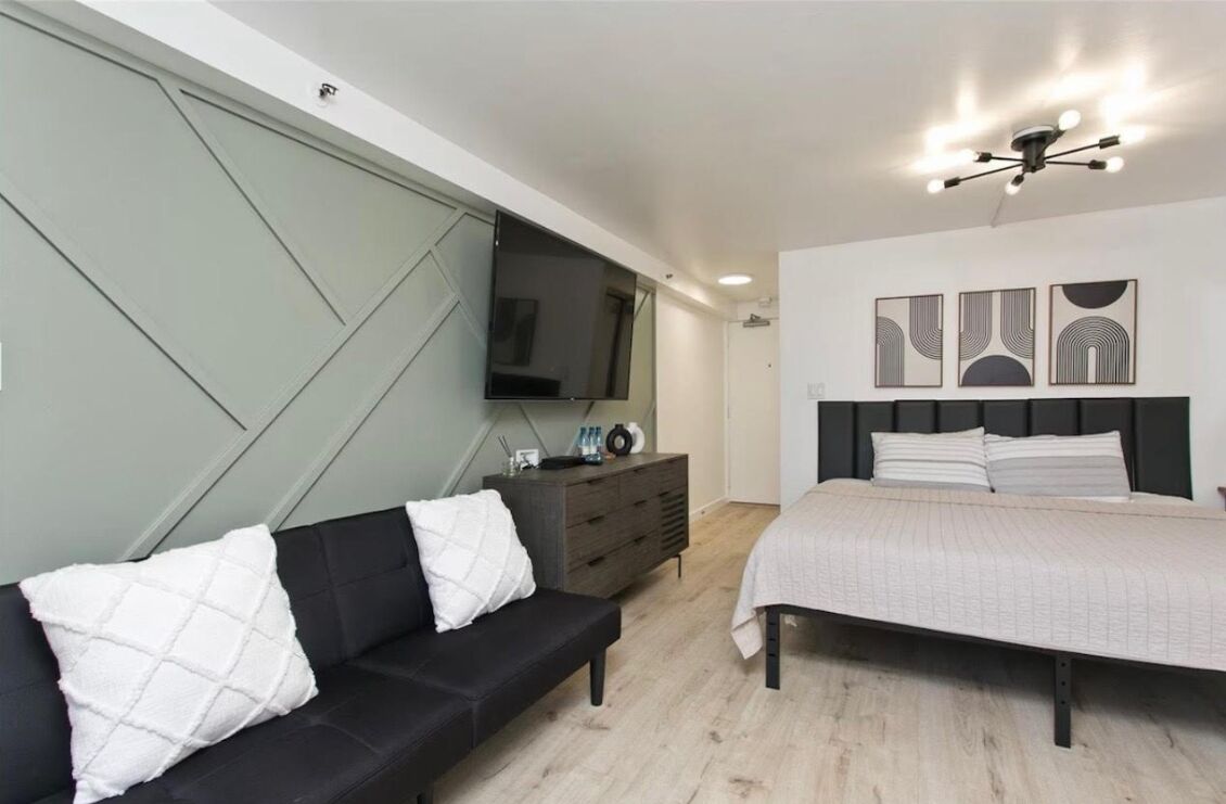 A modern bedroom with a bed, a black sofa, a TV mounted on the wall, and a ceiling light fixture.