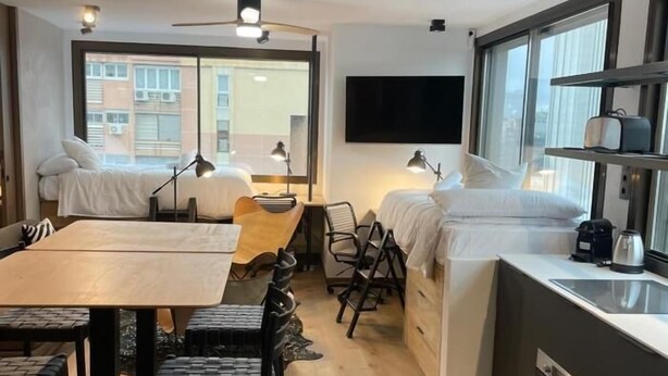 A compact hotel room with a bed, a desk, a chair, a small kitchenette, and a TV mounted on the wall. - Chic Studio Loft with City Views (Madrid)