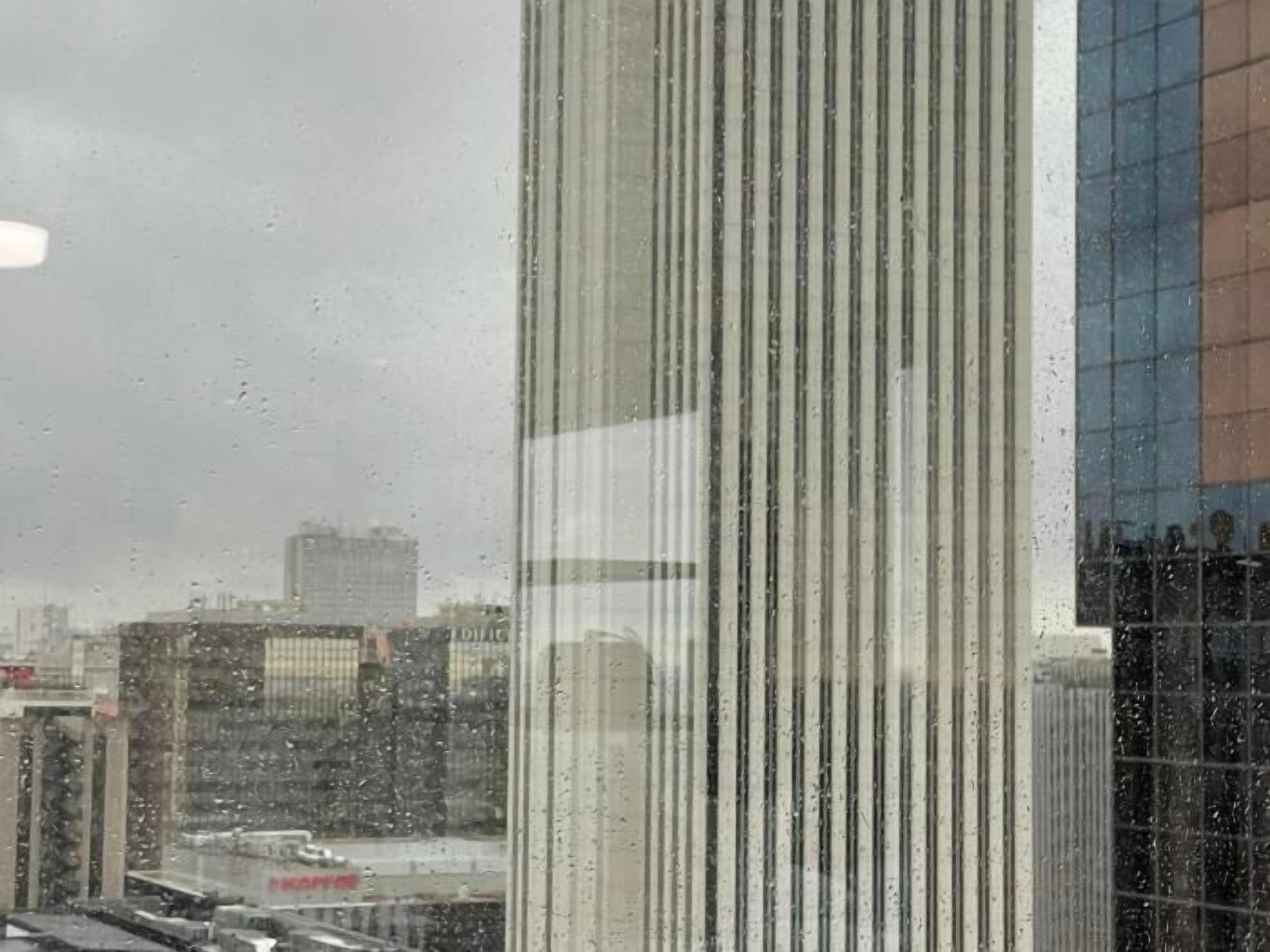 A view through a window with vertical blinds, a cityscape with buildings, and a reflection of a building with a blue and orange facade.