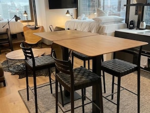 A modern hotel room with a wooden dining table, black upholstered chairs, a bed with white linens, a flat-screen TV mounted on the wall, and a window with a city view.
