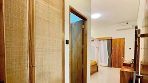 A room with a wooden wardrobe, a bed with a white duvet, a small table with a plant, and a wooden cabinet. - Lebak Bali Batu Mejan (Canggu)