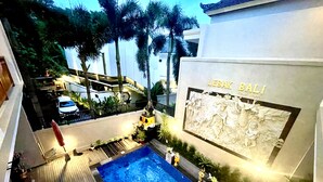 A swimming pool with a wooden deck, surrounded by palm trees and a building with a relief sculpture. - Lebak Bali Batu Mejan (Canggu)