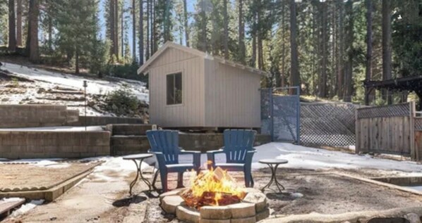 A small wooden cabin with a fire pit and two chairs in a forested area.
