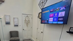 Un salón amplio con un televisor de pantalla plana grande montado en la pared, un sillón gris y suelo de madera.