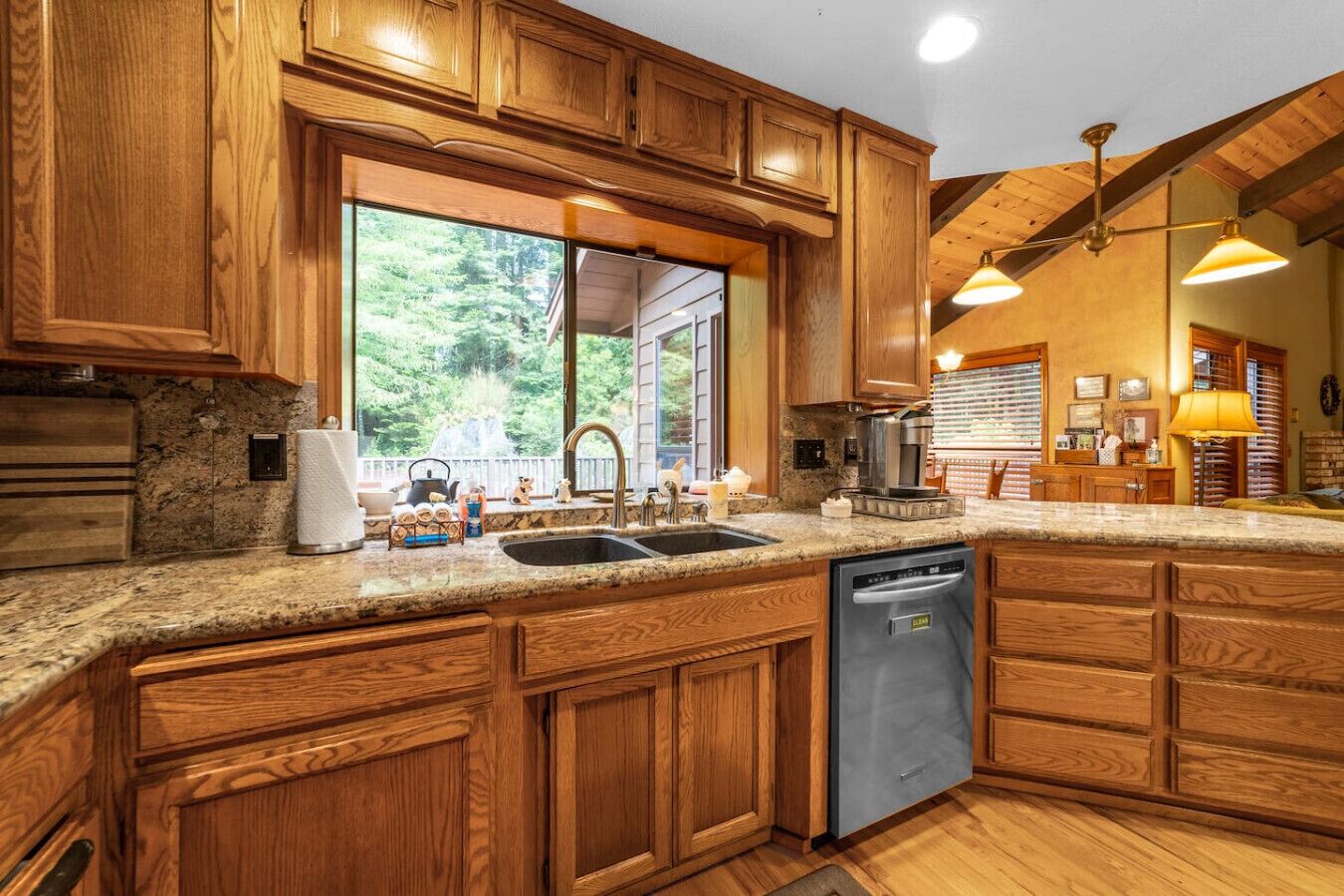 A kitchen with wooden cabinets, a granite countertop, a dishwasher, and a window overlooking a forest.