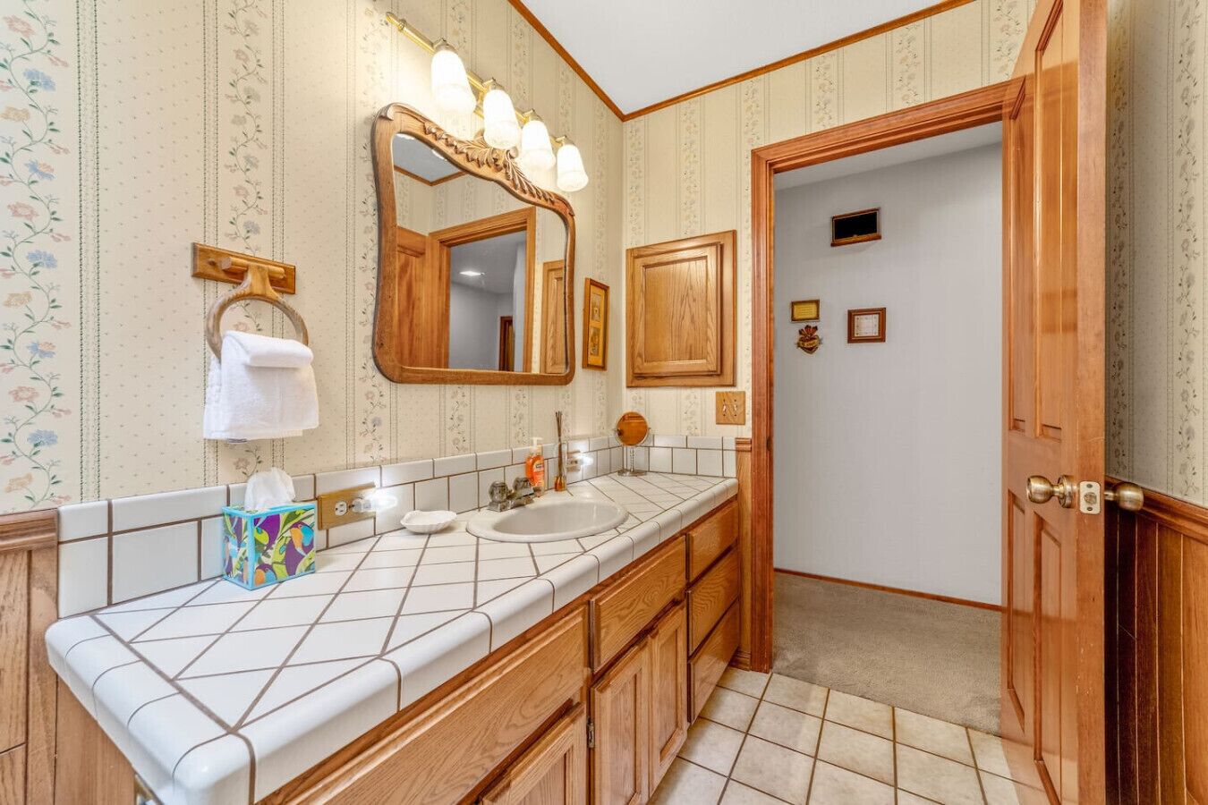A bathroom with a white tiled vanity, wooden cabinets, a large mirror with lights, and a door leading to another room.