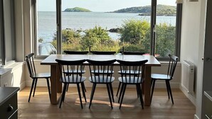 Un espace repas comprenant une table et des chaises, de grandes fenêtres offrant une vue sur la mer et la verdure.