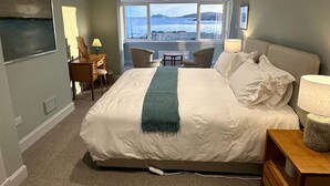 Une chambre d’hôtel avec un grand lit, un bureau avec une chaise, une petite table et une vue sur la mer par une grande fenêtre.