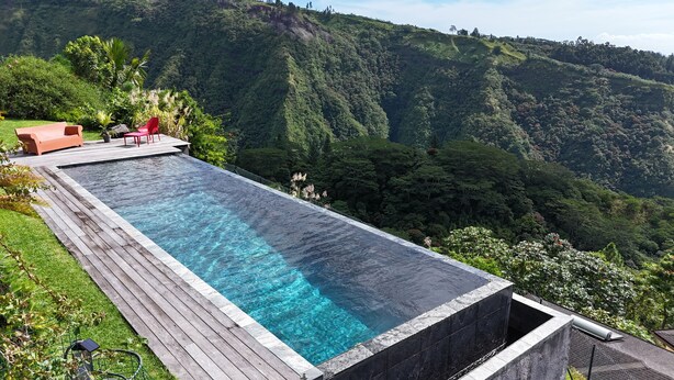 Una piscina infinita rectangular con cubierta de madera, rodeada de exuberante vegetación y un telón de fondo montañoso.