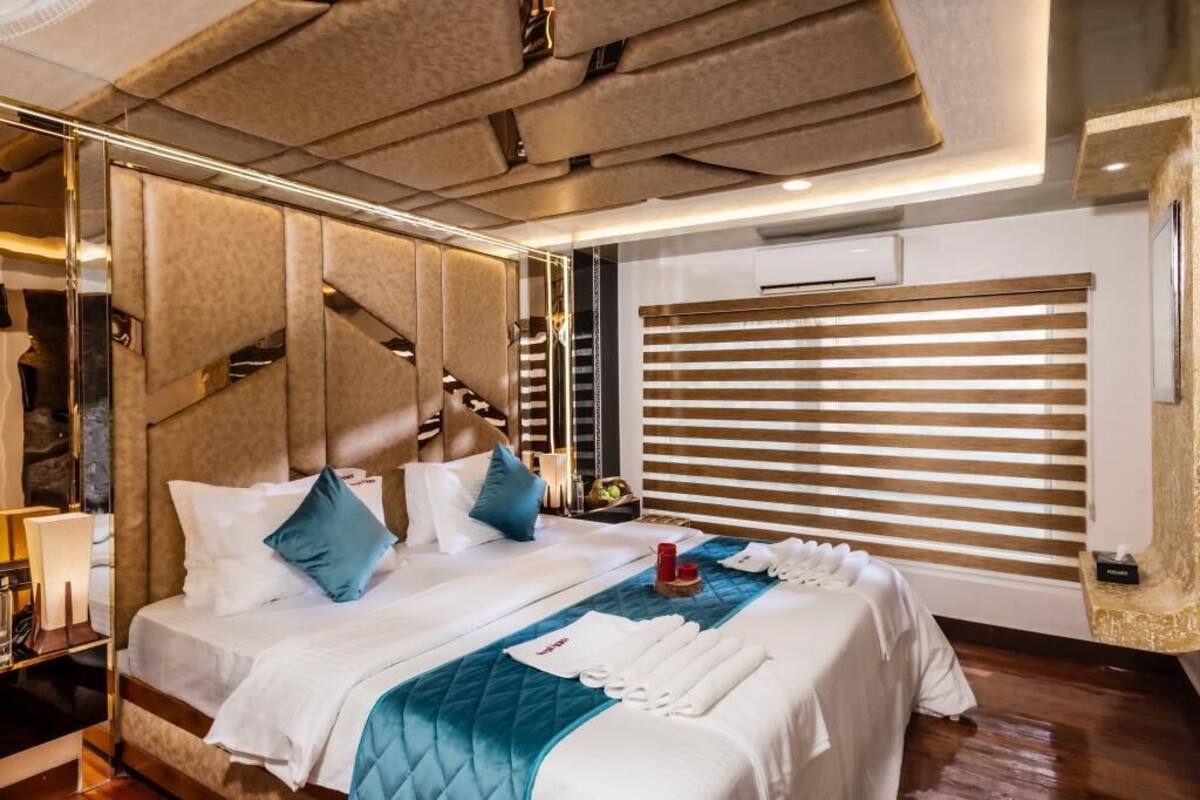 Een hotelkamer met een groot bed, een hoofdbord met een geometrisch patroon, een nachtkastje met een lamp en een raam met jaloezieën.