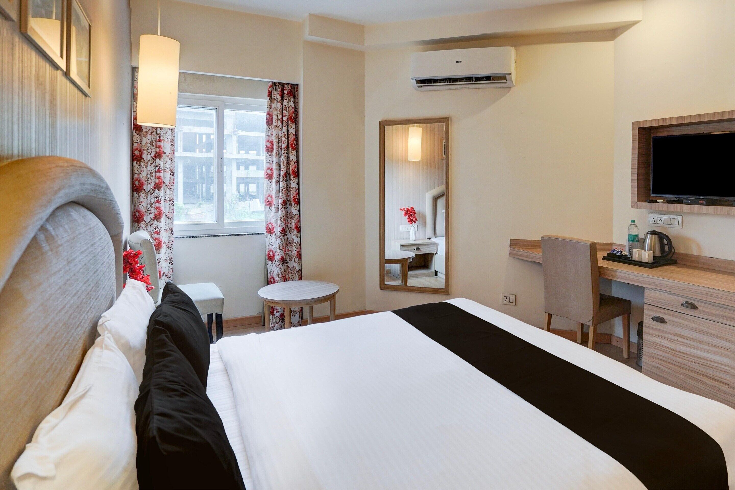 Et hotelværelse med seng, skrivebord, stol, lille bord, spejl og fjernsyn.