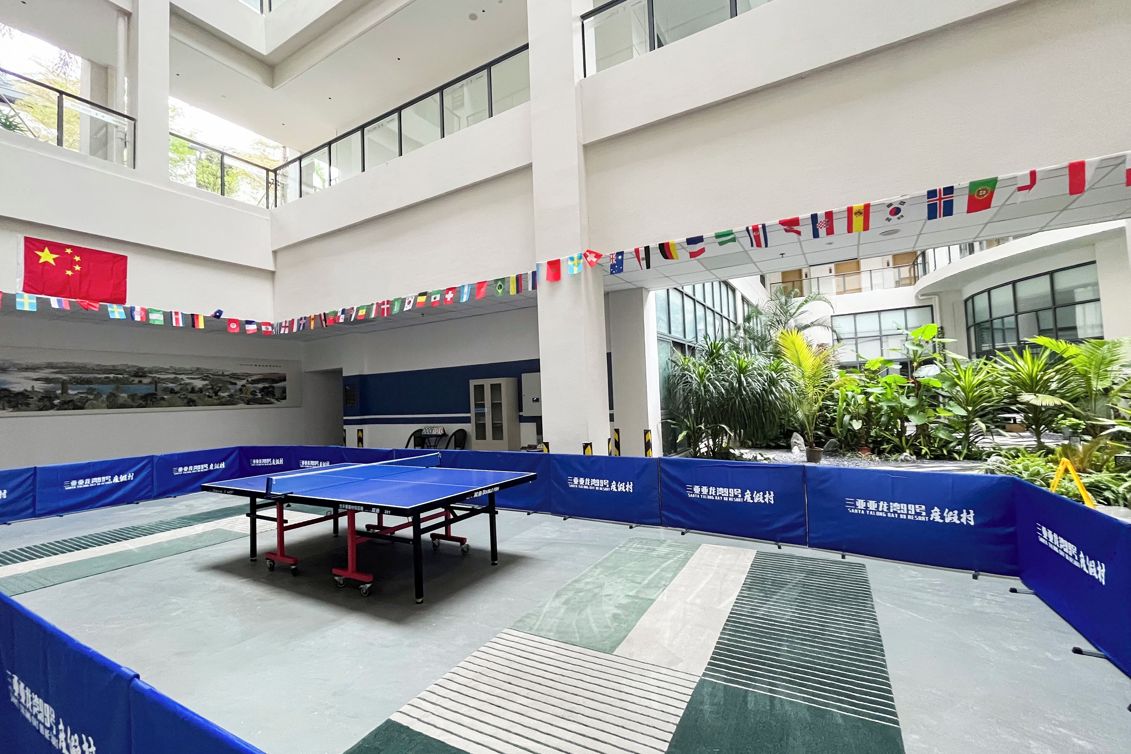 Una mesa de ping pong en un amplio espacio interior con varios niveles, rodeada por barreras azules y banderas internacionales.
