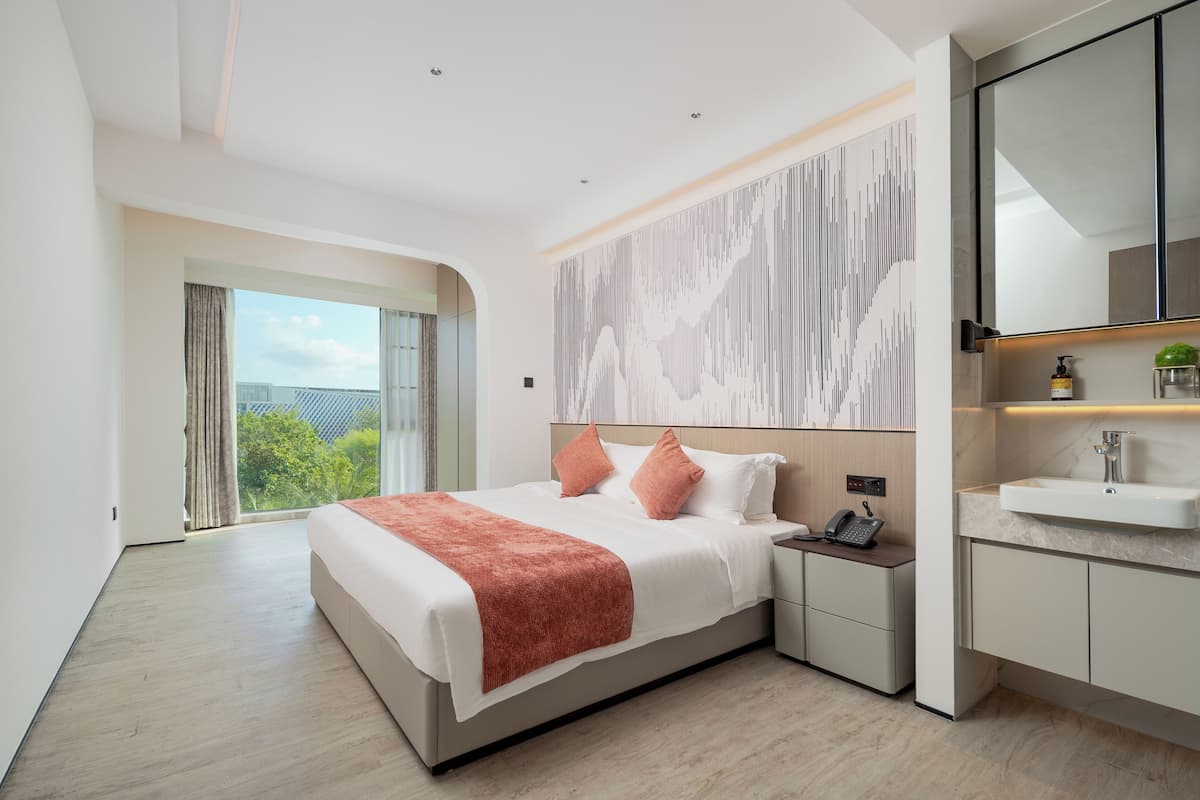 A modern hotel room with a large bed, a bedside table with a phone, a nightstand with a lamp, a bathroom with a sink, and a large window with a view of greenery.