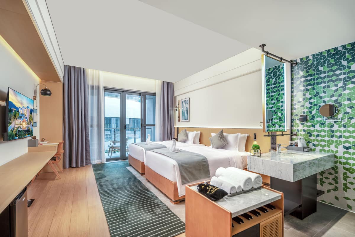 A hotel room with two beds, a desk, a large window, a bathroom with a green-tiled wall, and a view of the city.