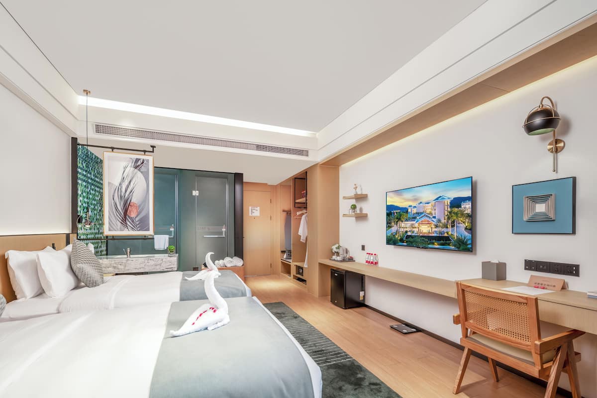 A modern hotel room with a large bed, a desk, and a chair. There is a painting on the wall and a window with a view.