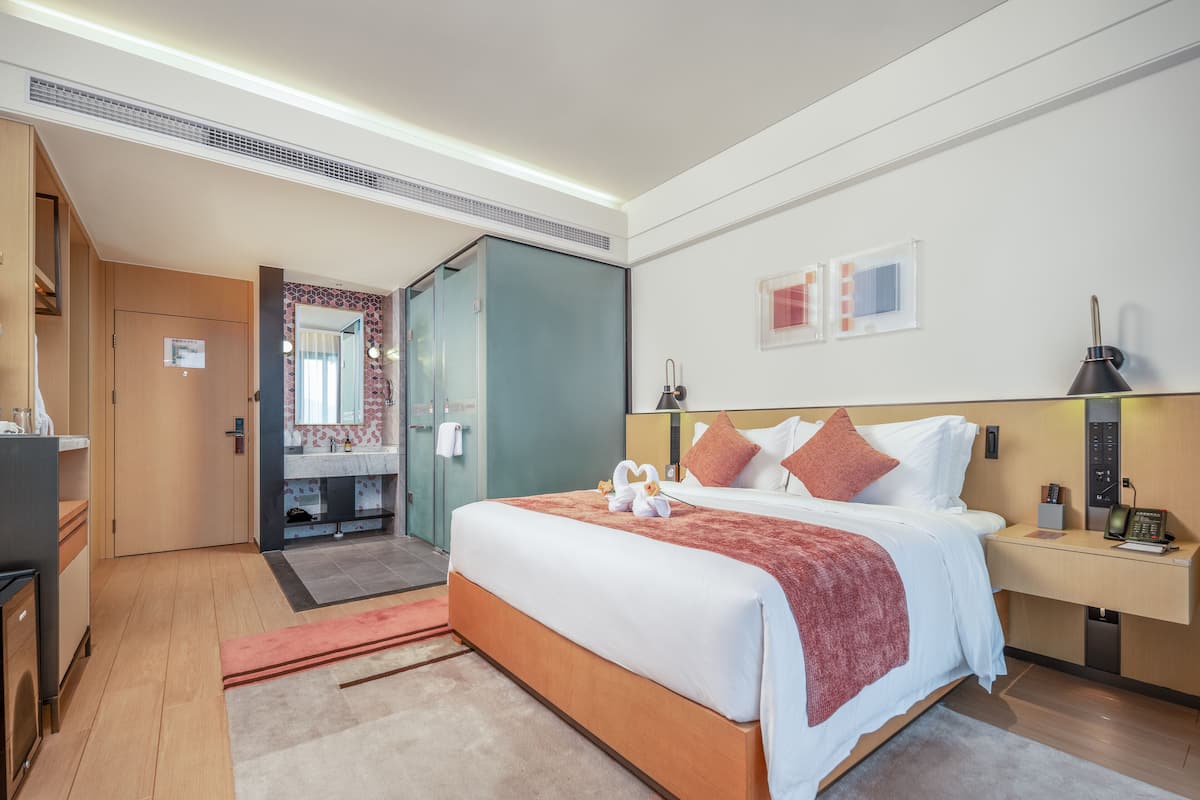 A modern hotel room with a large bed, a desk, a mirror, and a bathroom visible through an open door.