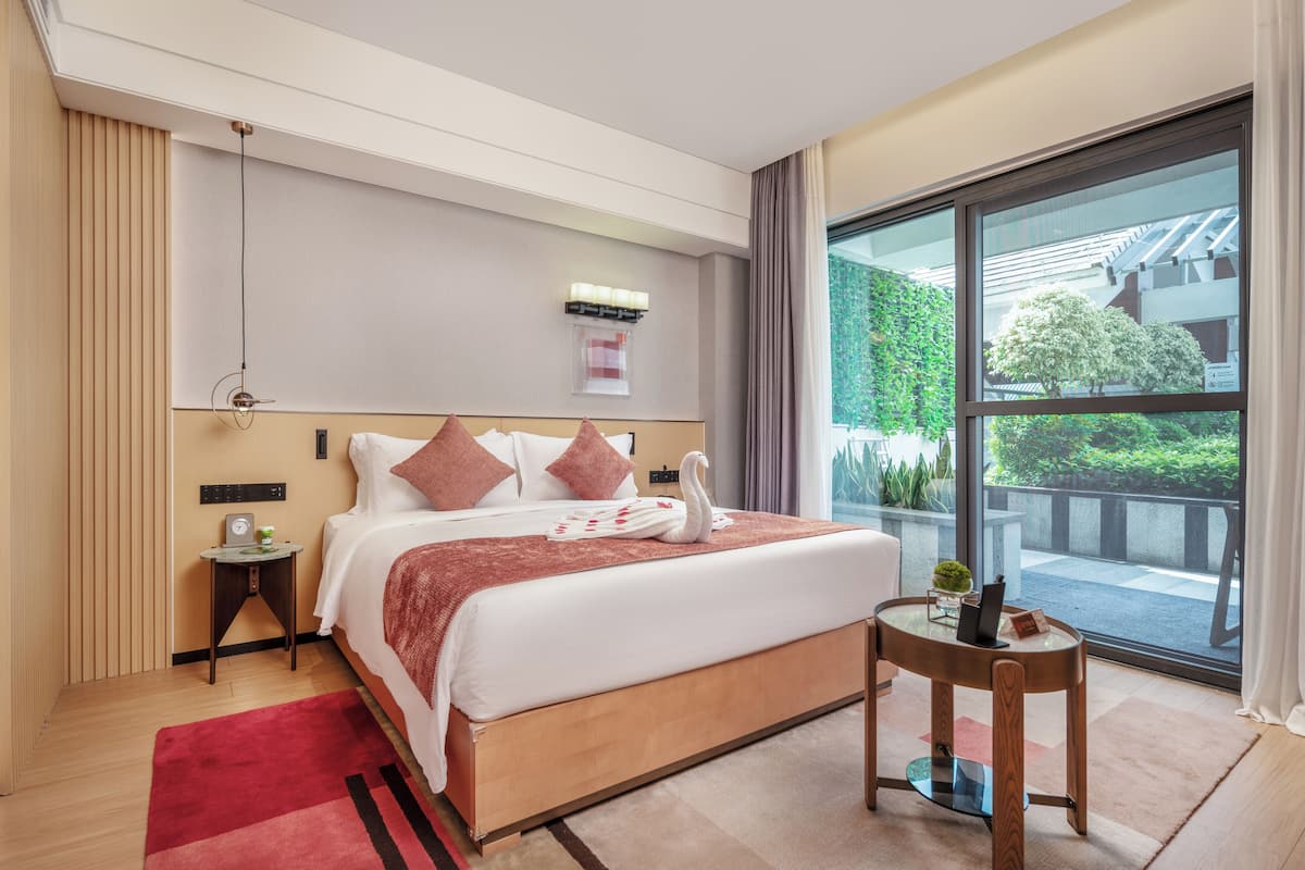 A hotel room with a large bed, a bedside table, a small table with a lamp, and a view of greenery outside.