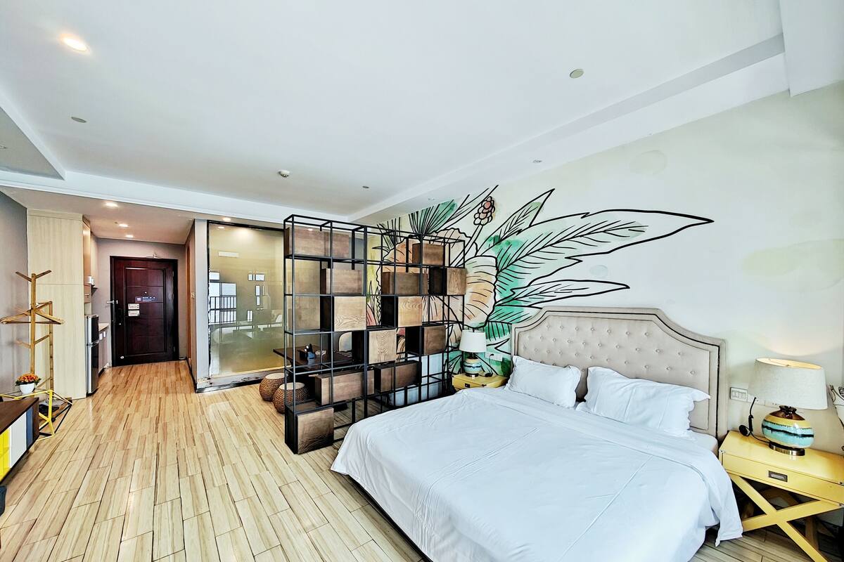 A hotel room with a large bed, a wooden floor, a decorative wall panel, and a view into the hallway.