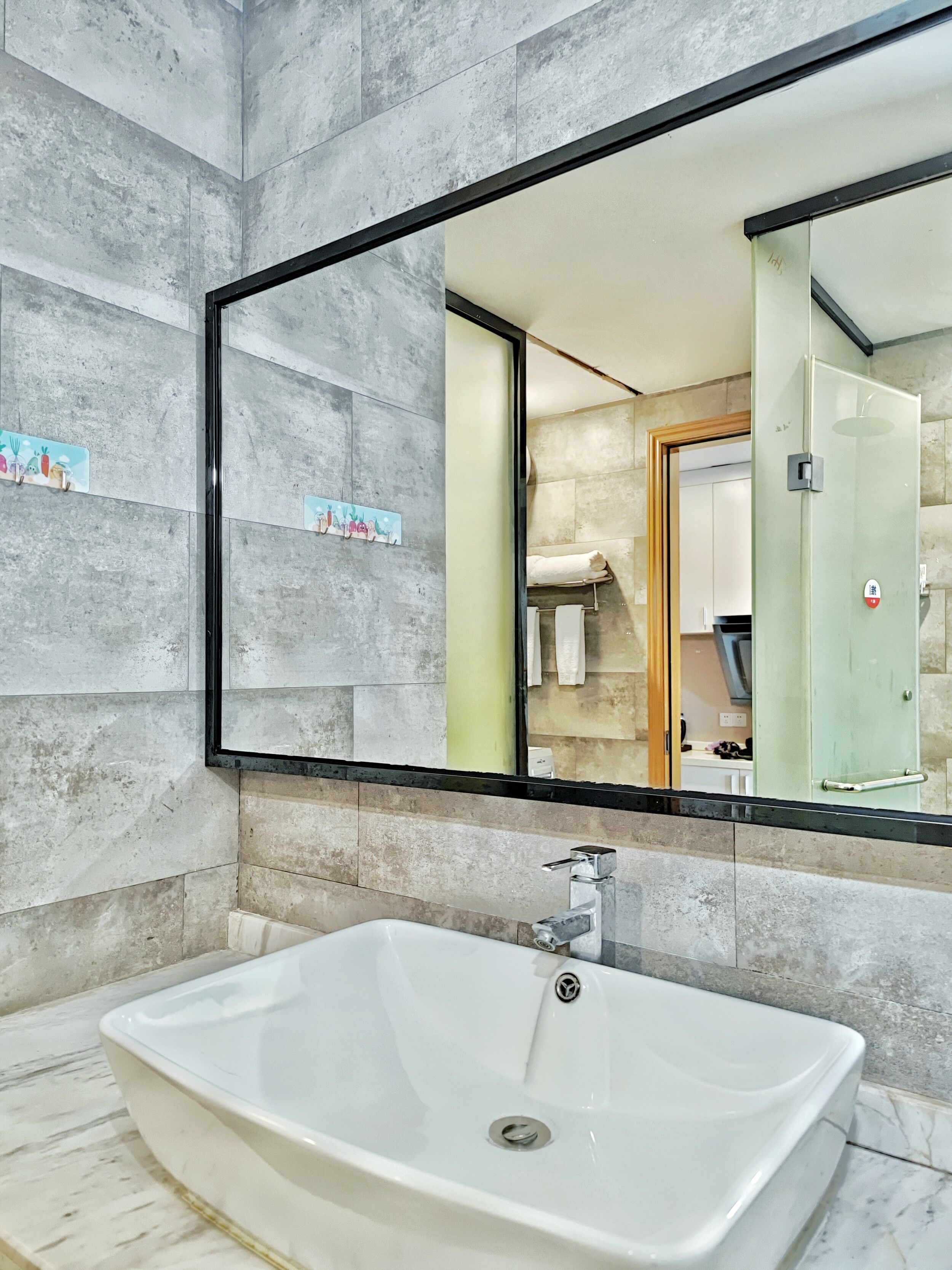 Ein modernes Badezimmer mit einem großen, rechteckigen Waschbecken, einem Spiegel mit schwarzem Rahmen und einer gefliesten Wand.