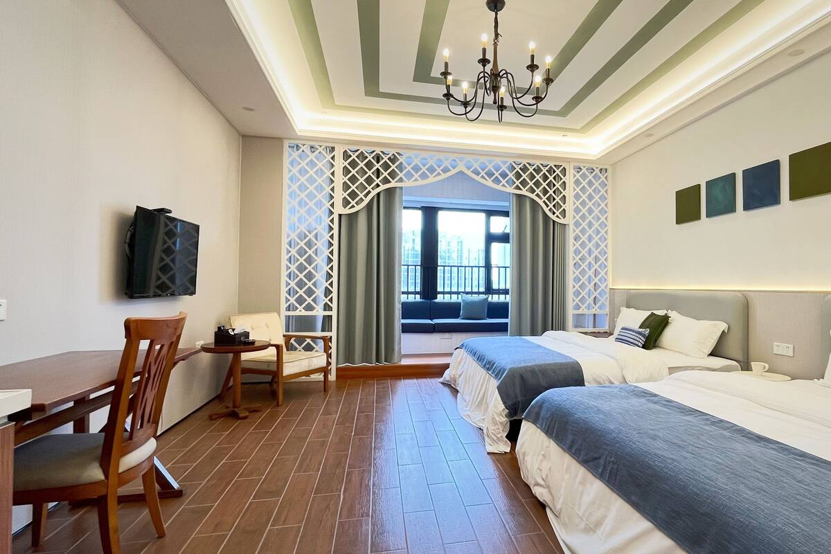 A hotel room with a wooden dining table, two beds, a TV mounted on the wall, and a view of the city through a window with a lattice design.