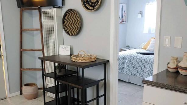 Una habitación pequeña y bien iluminada con televisión, una mesa con una cesta, una escalera y una puerta que da a otra habitación con cama.