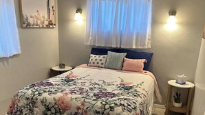 Un dormitorio con colcha estampada, una mesita de noche con lámpara y un equipo de aire acondicionado sobre la ventana.