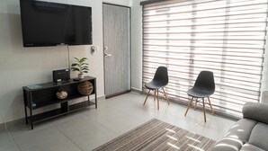 Een moderne woonkamer met een flatscreen televisie, een bank, twee fauteuils, een console met een plant, en een schuifpui met zonwering.