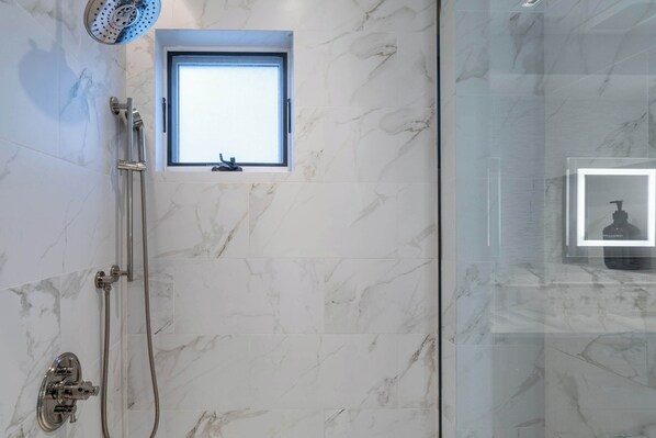 Une salle de bain moderne avec des carreaux de marbre, une douche équipée d’un pommeau de douche à main, une fenêtre et un espace pour les toilettes séparé par une paroi de verre.