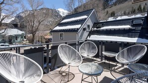 A balcony with white wire chairs and a table, overlooking a snowy mountain and a house with solar panels.
