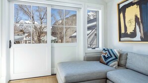 A room with a sliding glass door leading to a snowy mountain view, a gray sofa with patterned cushions, and a large abstract painting on the wall.