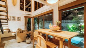 A spacious living area with a wooden dining table, comfortable seating, a staircase, and large windows offering views of the outdoors. - Villa Jamu Bingin (Pecatu)