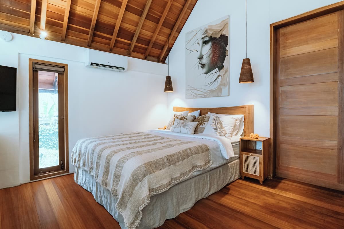 Een slaapkamer met een houten plafond, een groot bed met gestreepte beddengoed, een houten nachtkastje en een wanddecoratie.