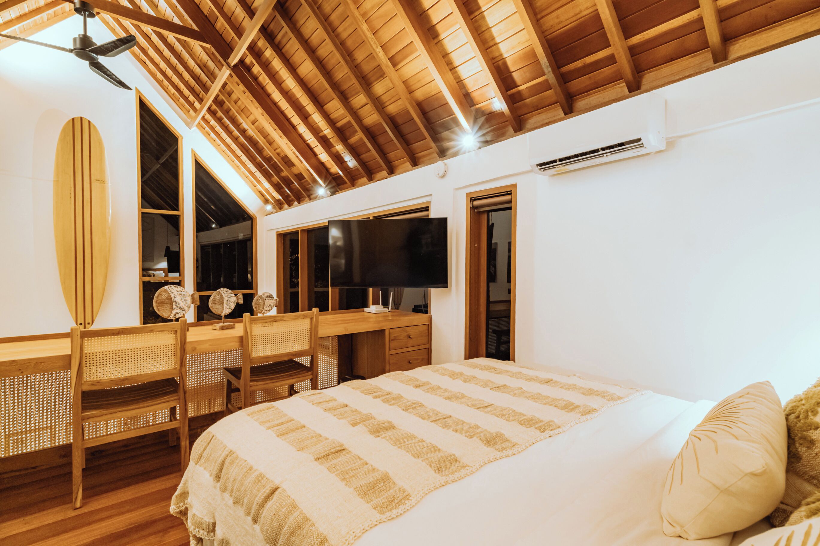 Un dormitorio con techo de madera, una cama con manta a rayas, un escritorio con computadora y una tabla de surf en la pared.