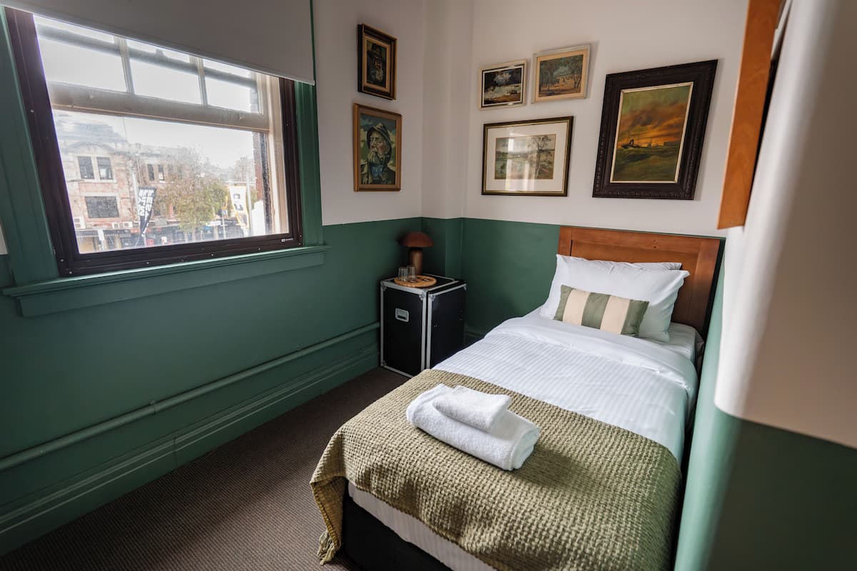 Quarto de hotel com uma cama, uma mesa de cabeceira, uma janela com vista para prédios e algumas fotografias emolduradas na parede.