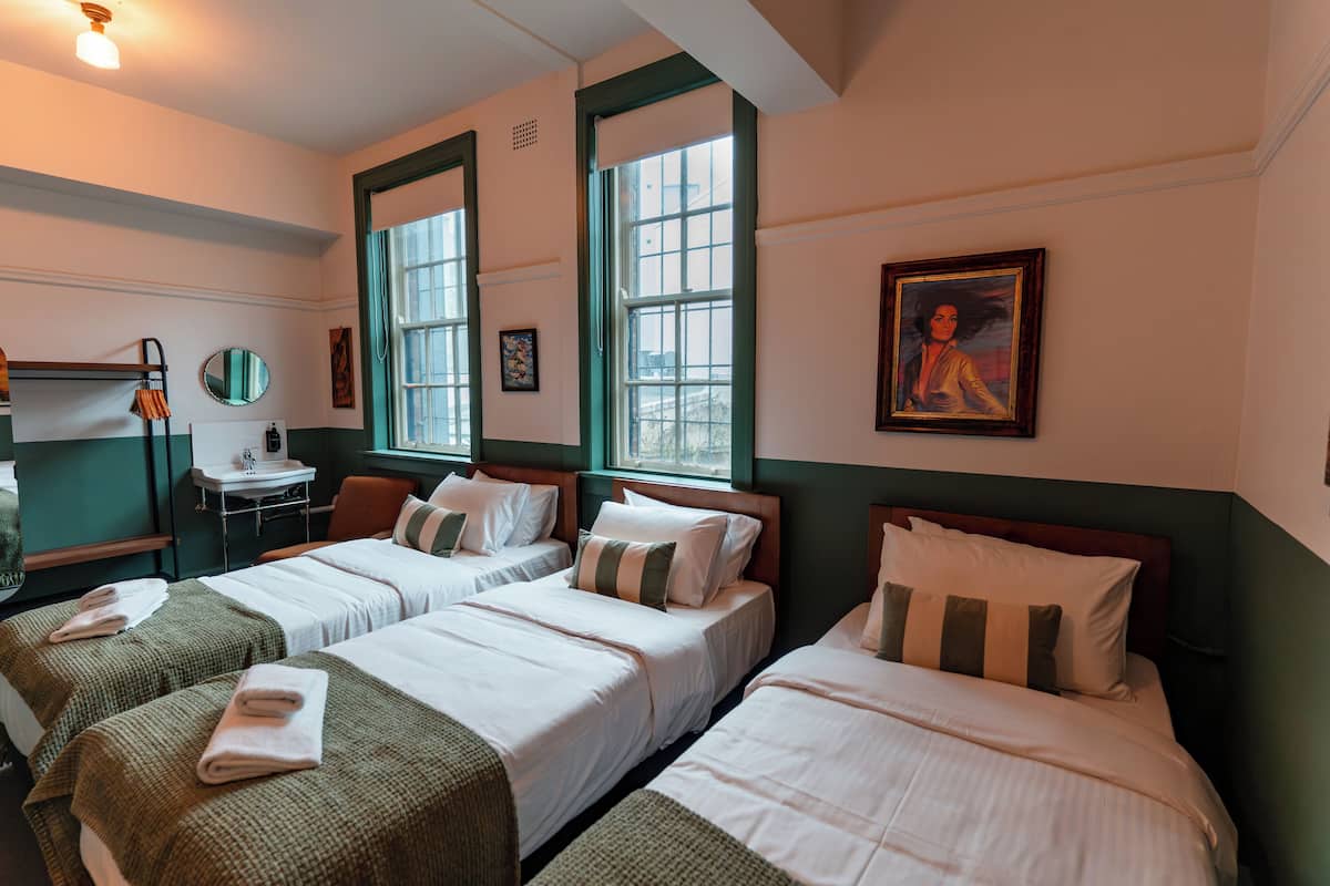 Quarto de hotel com duas camas de solteiro, uma escrivaninha com cadeira, um espelho e uma janela.