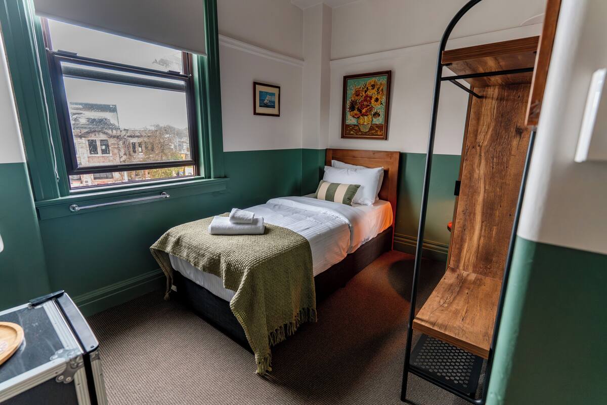 Um quarto com uma cama, uma janela, um banco de madeira e uma pequena mesa.