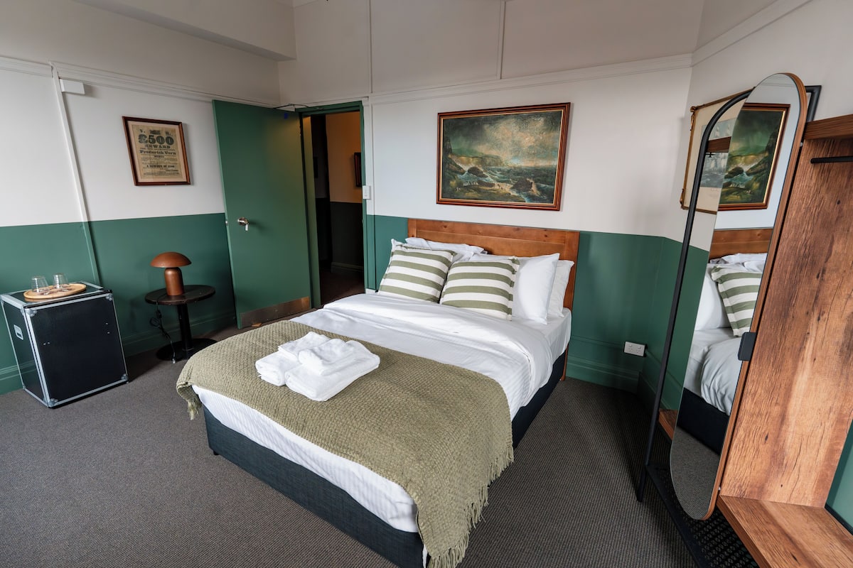 Quarto de hotel com cama, abajur, mesinha e espelho.
