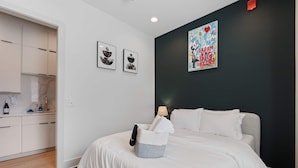 Un dormitorio con cama, mesita de noche con lámpara, pared con cuadro y un rincón de kitchenette.