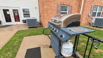 An outdoor grill setup with a propane tank, positioned in front of a brick building with a door and windows.