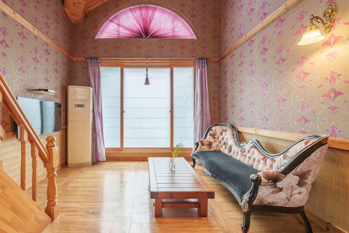 Ein gemütliches Wohnzimmer mit einer Holztreppe, einem Sofa, einem Couchtisch und einem großen Fenster mit Vorhängen.