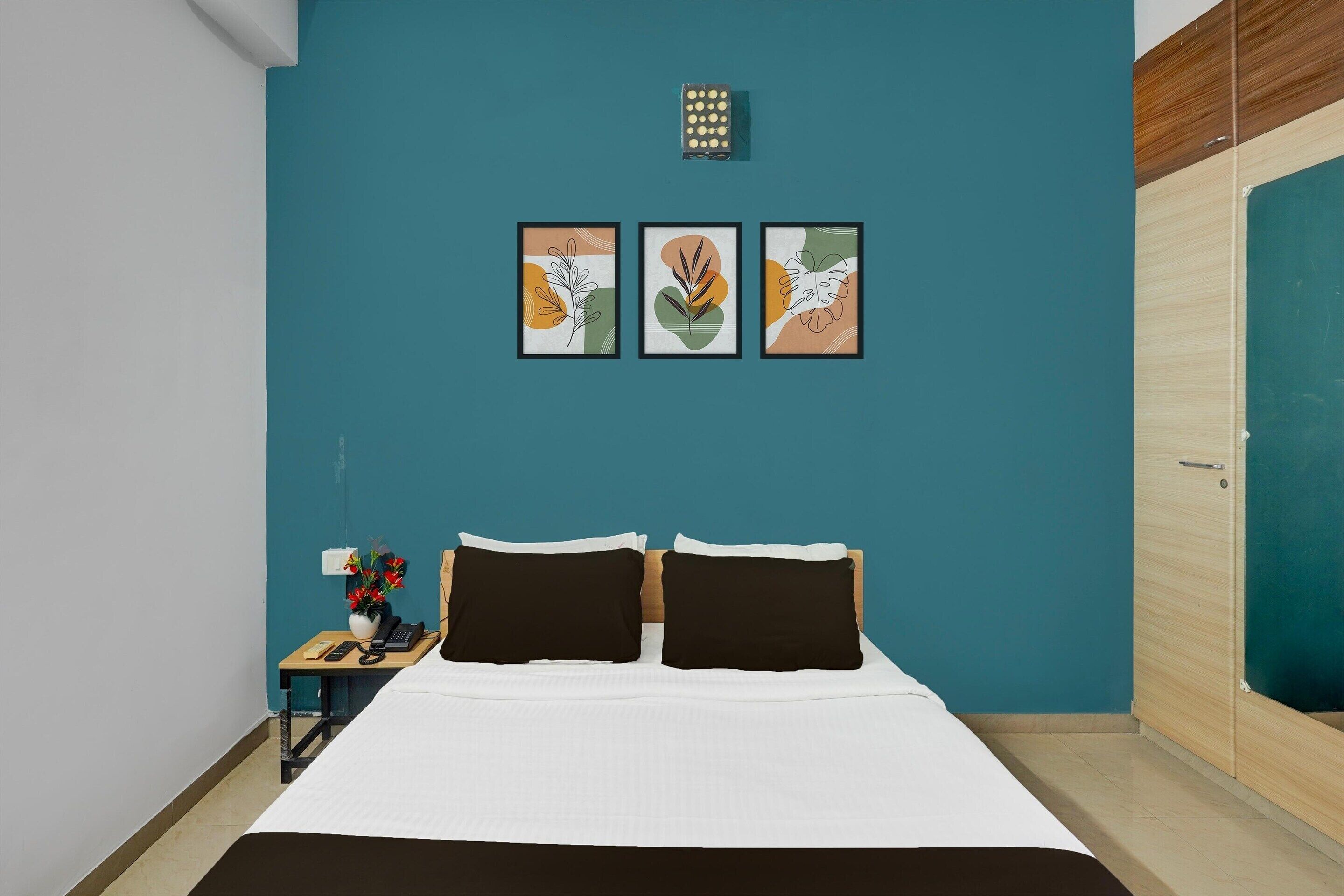 A bedroom with a teal wall, a bed with white and brown bedding, two black pillows, a nightstand with a vase of flowers, and three framed artworks above the bed.