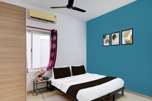 A bedroom with a bed, a bedside table with a phone and a vase of flowers, a window with curtains, an air conditioner, and three framed pictures on the wall.