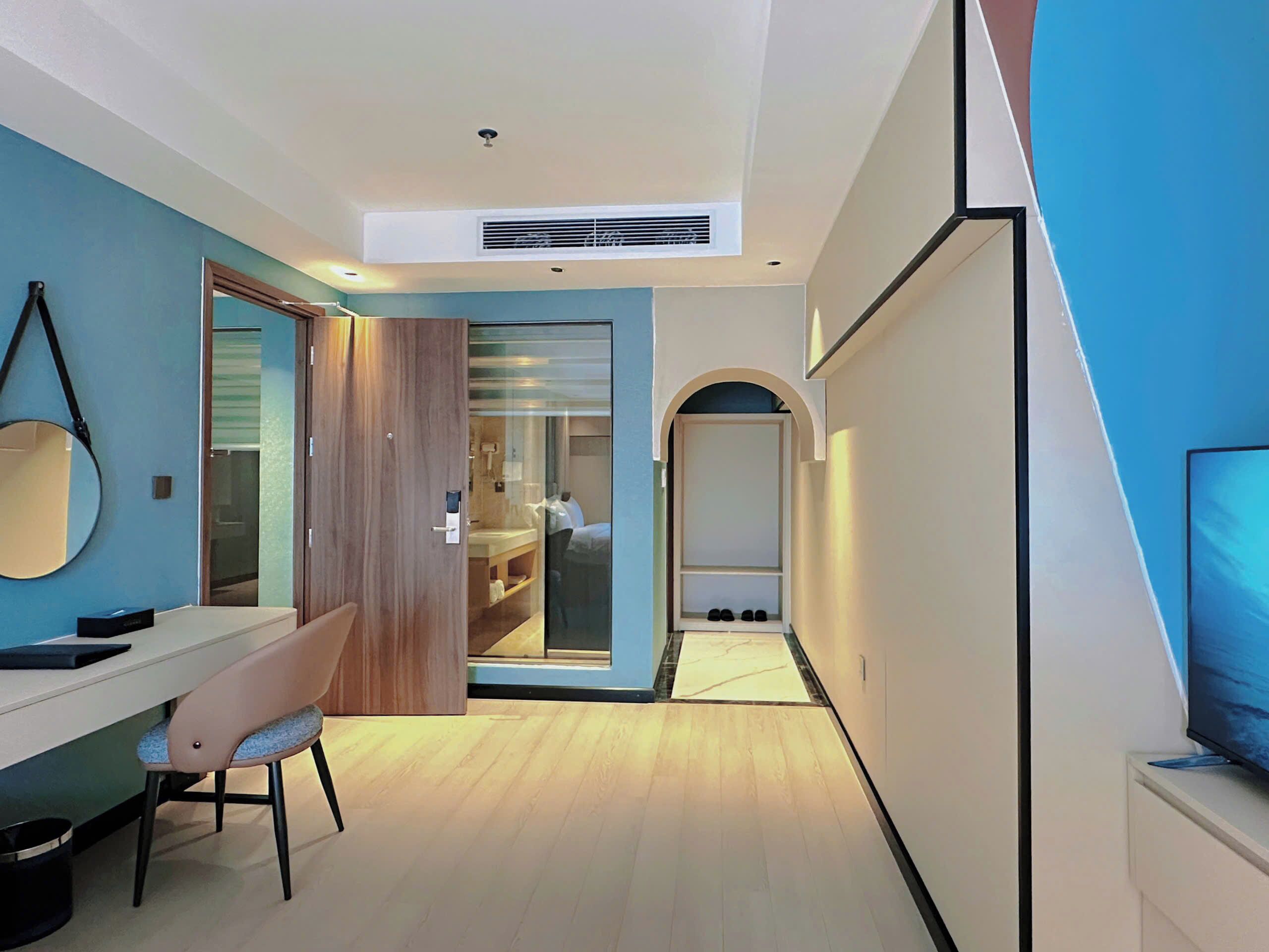 A modern room with a desk, chair, and a large mirror. There is a door leading to a bathroom with a shower and a small window.
